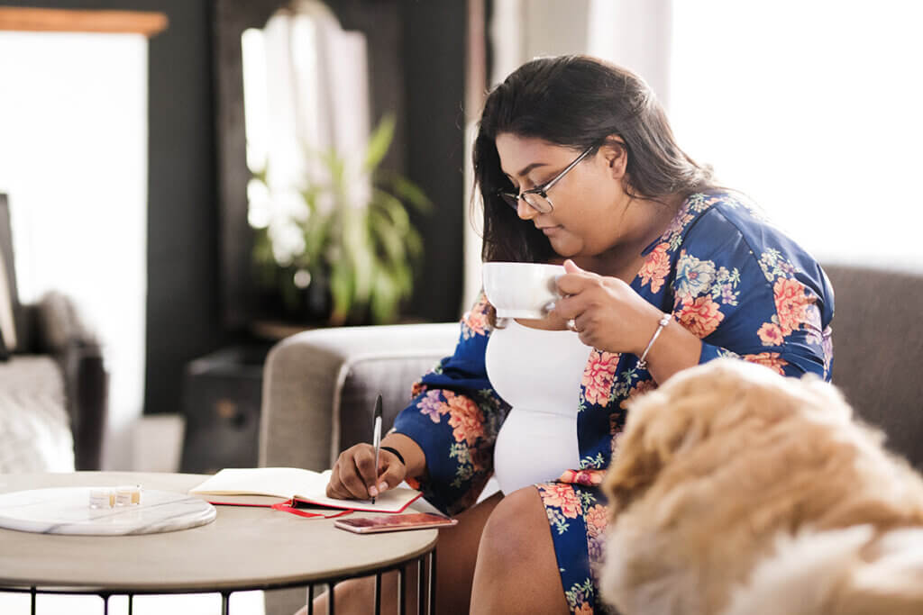 Shot of a woman writing in her diary while sitting in living room with a cup of coffee